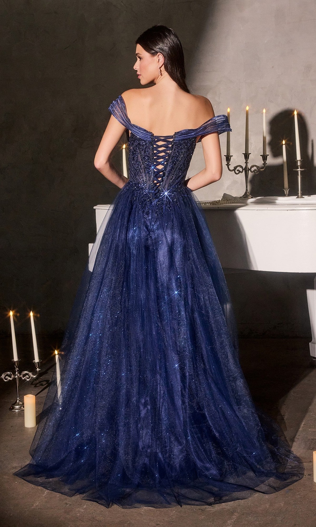 A woman wearing the Ladivine CC493 Long Prom Dress in dark blue, featuring an off-shoulder sweetheart neckline, stands facing away before a white piano and glowing candles.